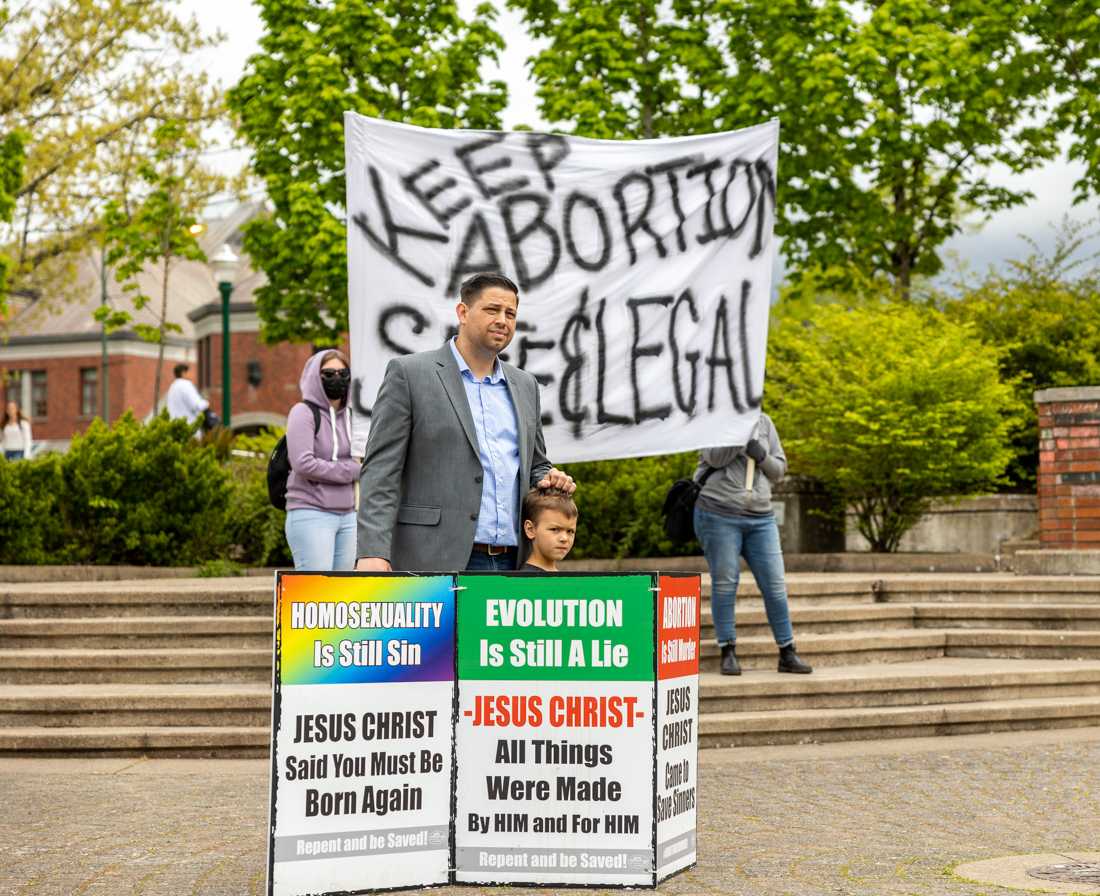 A man stands with a young boy and a sign in the middle of the amphitheater as counter-protesters stand behind them. Anti-abortion protestors are met with counter-protesters outside the Erb Memorial Union on May 5th, 2023. (Molly McPherson/Emerald)