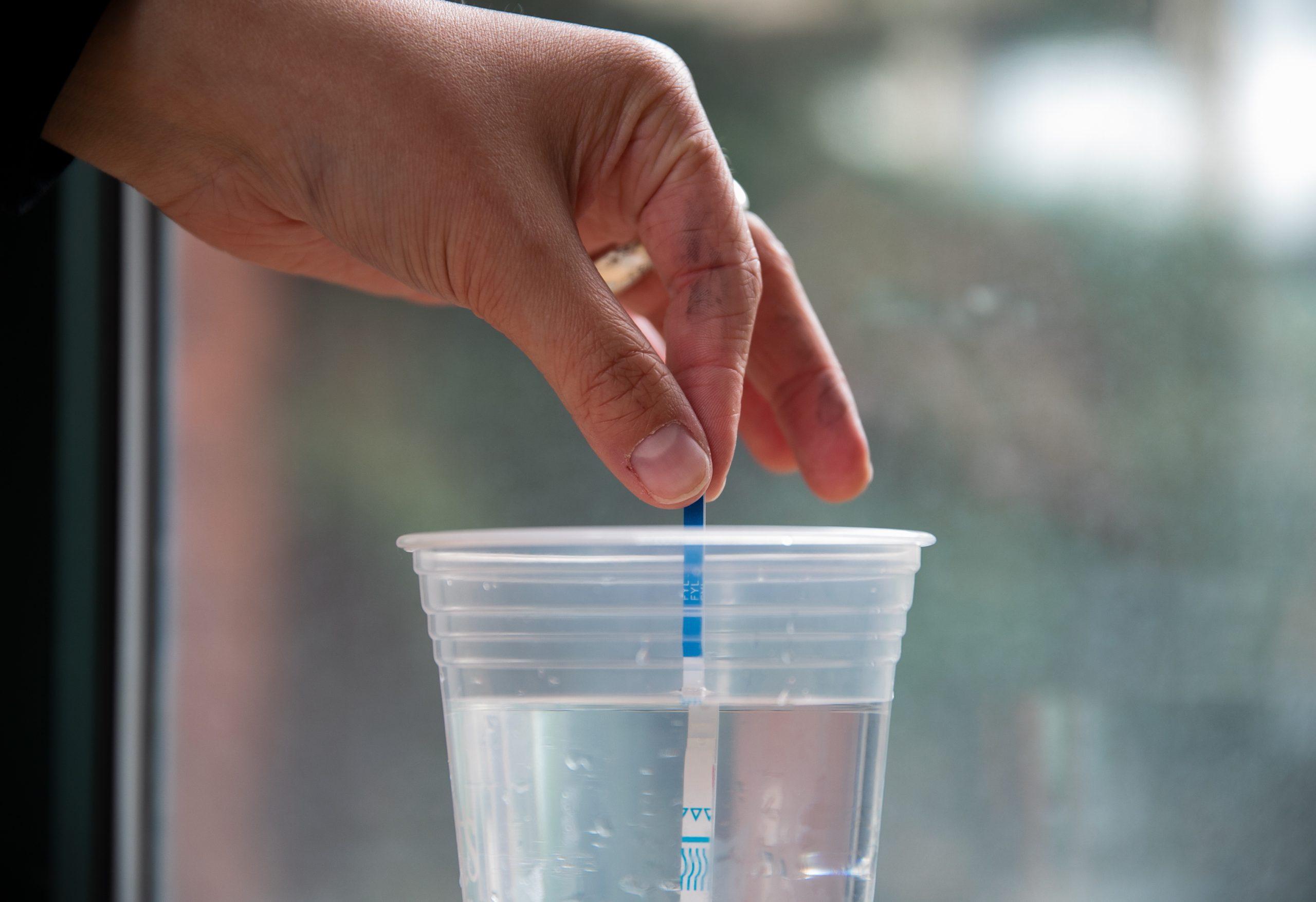 ASUO member Kayla Krueger&#160;demonstrates how to use a test strip after dissolving the substance in a cup of water. (Liam Sherry/Emerald)