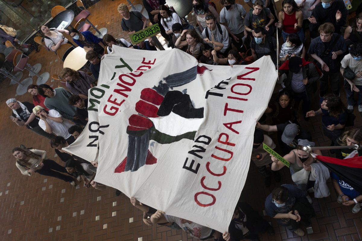 Protesters fill the Lillis Business Complex atrium and hold a banner that reads, "No Money For Genocide" and "End the Occupation." Protesters gathered on May 20, 2024, for a "100 Hours in Chains" rally in front of Johnson Hall&#8212;now "renamed" by protesters to "Alareer Hall," after writer and poet Refaat Alareer, who was killed in Gaza&#8212;with students who had chained themselves to the building removing their chains before marching through other buildings on campus; the rally is the most recent in a series of pro-Palestine demonstrations on the UO campus. (Colleen Bogdan/Emerald)