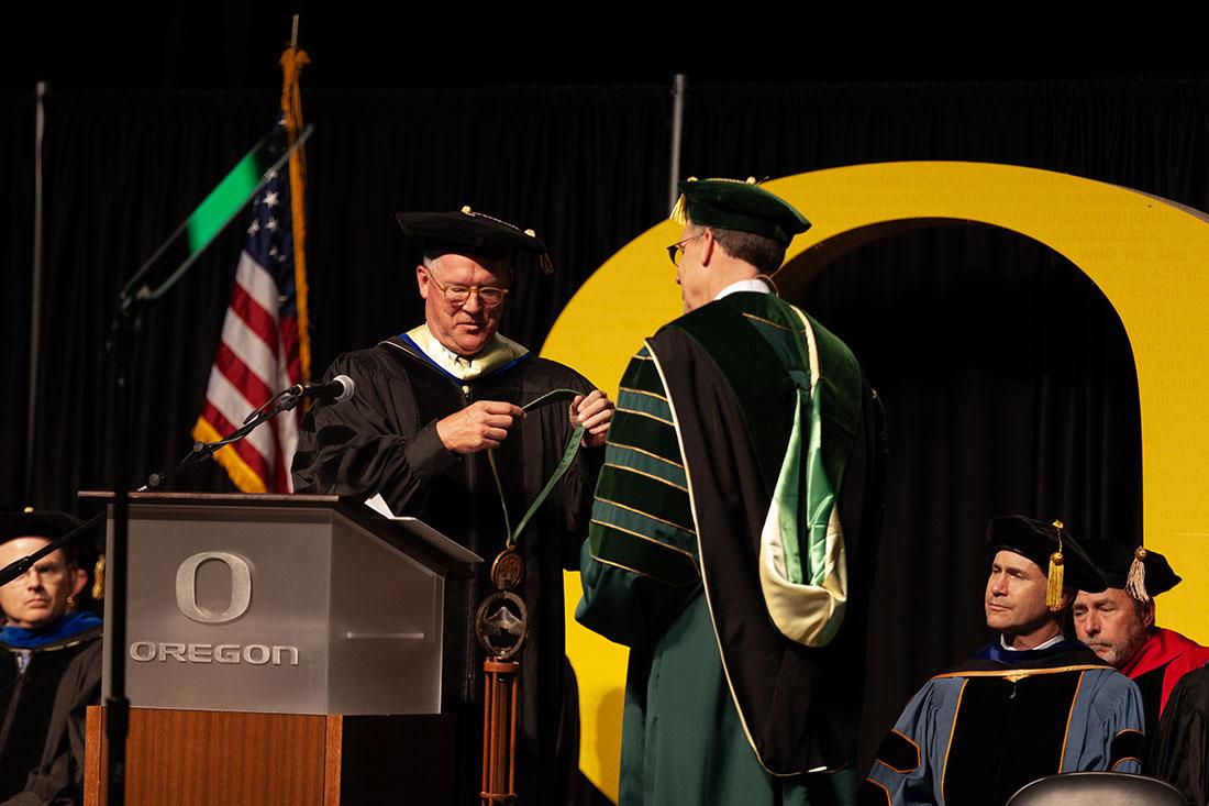 Steve Holwerda passes the Centennial Medallion to Karl Scholz. Thursday, President of the University of Oregon, Karl Scholz, attended his investiture at the Matthew Knight Arena, celebrating his formal installment as president of the university. Pro-Palestine protestors interrupted the ceremony as Scholz accepted the Centennial Medallion with a deluge of chants. After a brief confrontation, protestors moved outside to a separate entrance where they continued their protest. (Miles Cull/Emerald)