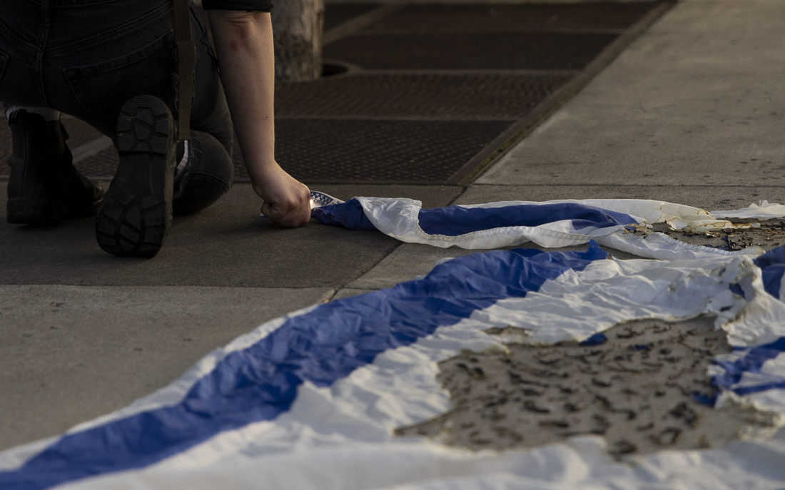 A demonstrator attempts to reignite the partially-burnt Israeli flag outside of the EMU as the march continues on Oct. 7, 2024. Protesters gathered for the anniversary of the Hamas-led attacks on Israel to protest the ongoing Israel-Hamas war and show solidarity with its victims. The march began at the Wayne Lyman Morse U.S. Courthouse and ended at the corner of E 18th Ave. and University St. (Alex Hernandez/Emerald)