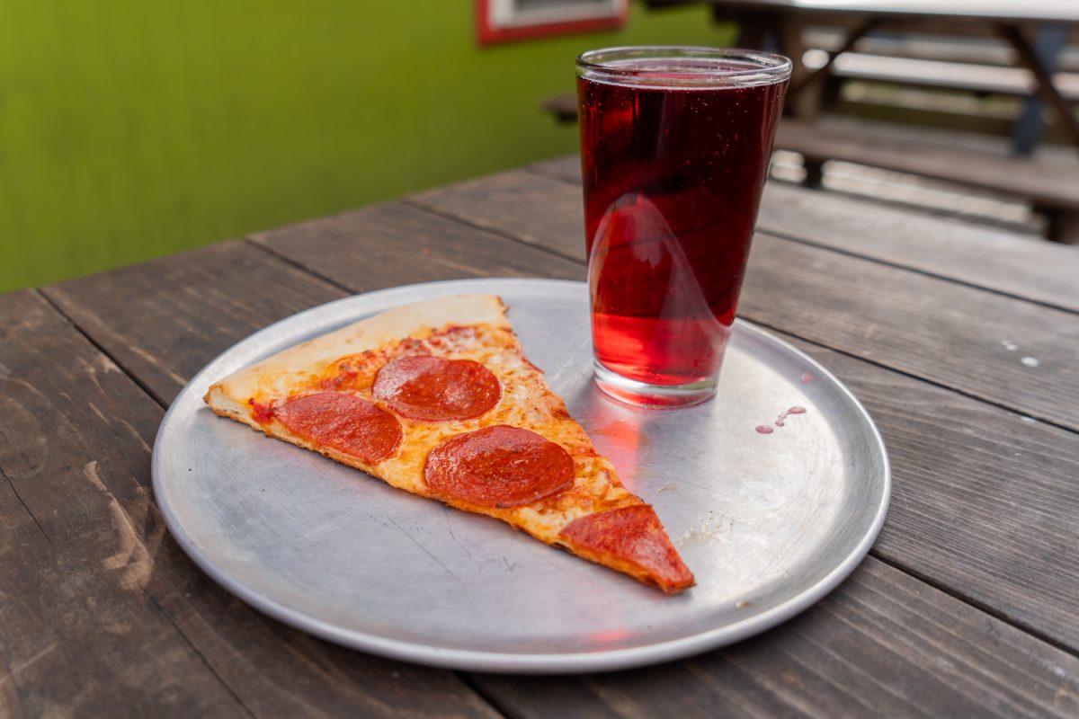 A slice of peperoni pizza and pint of raspberry cider on the Pegasus Pizza deck. The pint and slice deal is offered wednesdays to students near the University of Oregon campus, photo taken on Mar. 17, 2026. (Rowan Campbell/Emerald).