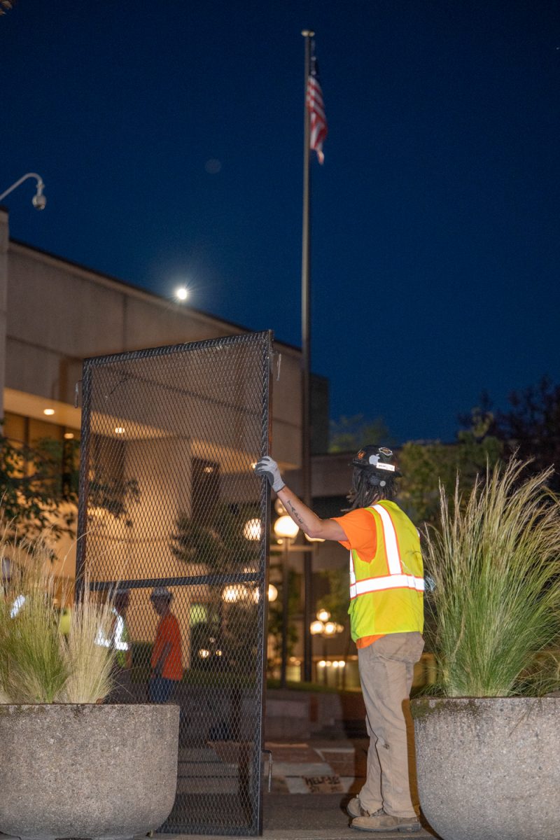 A worker waits with a section of fence as the American flag flies above him, on the property of the Federal Building. A fence surrounding the Federal Building in Eugene, Ore., is removed on July 1, 2026. The fence, which had been erected on the property after a series of protests, was ordered to be removed by a judge after it was found to violate the First Amendment rights of people wishing to assemble there. (Chris Castro/Emerald)